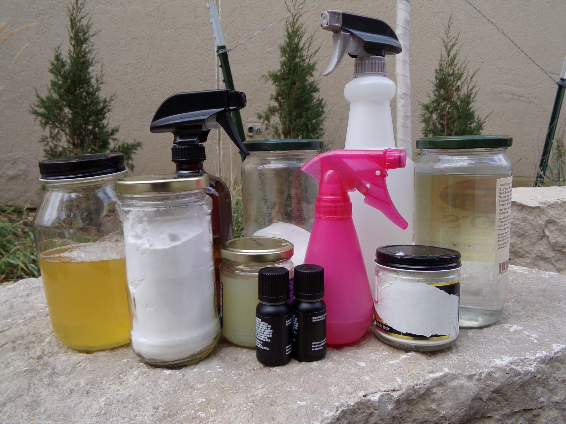 A glass jar of castile soap, a glass jar of baking soda, a glass jar of washing soda, an amber glass trigger spray bottle of cleaning fluid, a small glass jar of lemon juice, two small glass bottles of essential oils, a small, pink, plastic trigger spray bottle of cleaning fluid, a small jar of corn starch, a tall, plastic, clear trigger spray bottle of cleaning fluid, and a glass jar of vinegar all sit together atop a rock.