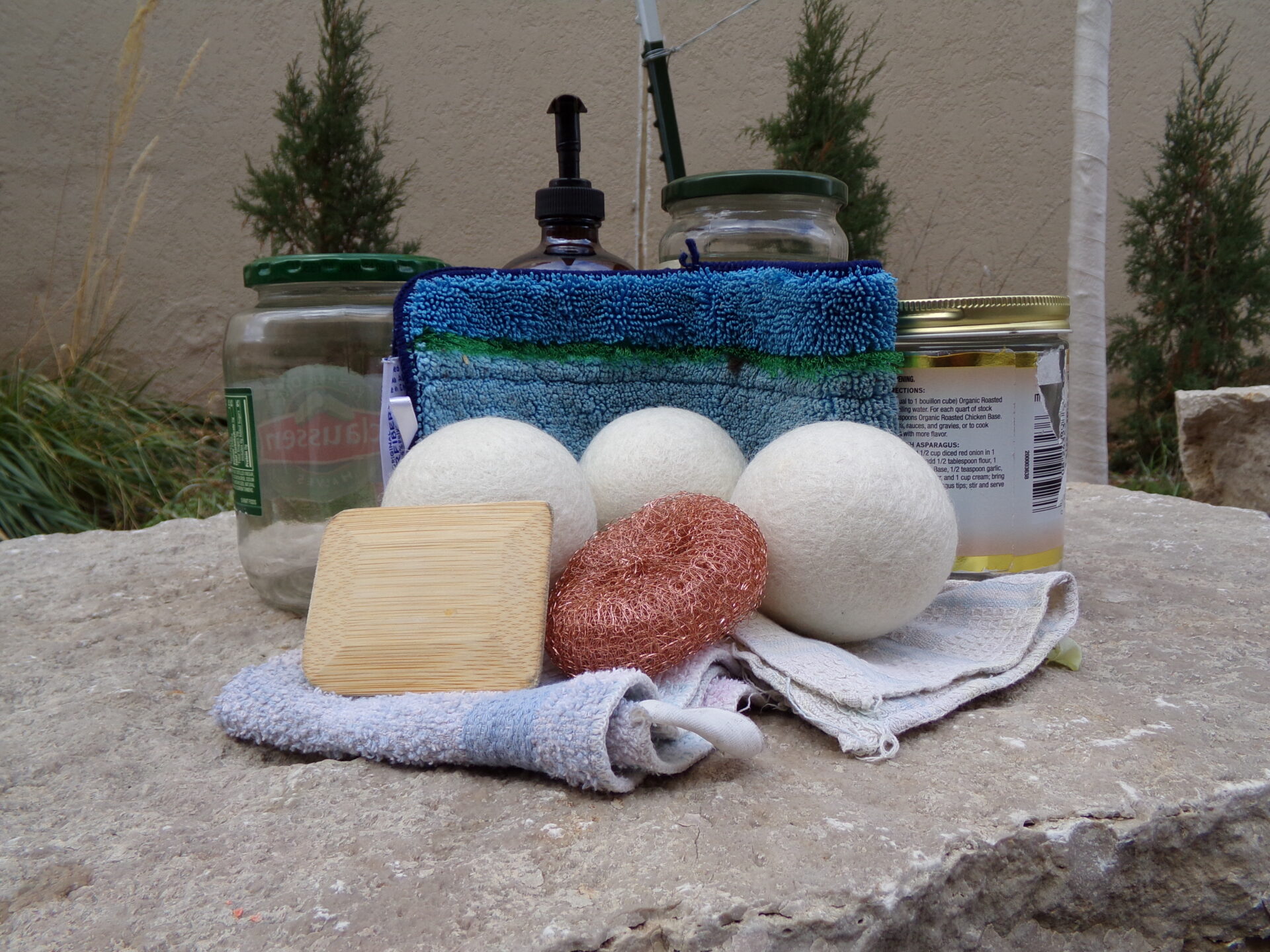 Three glass jars, a glass pump bottle, three wool balls, two rags, a copper scrubber, a bamboo scraping tool, and a blue washable mop pad all sit together atop a rock.